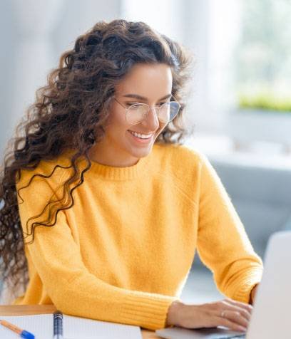 a woman in a yellow sweater and glasses typing on a laptop